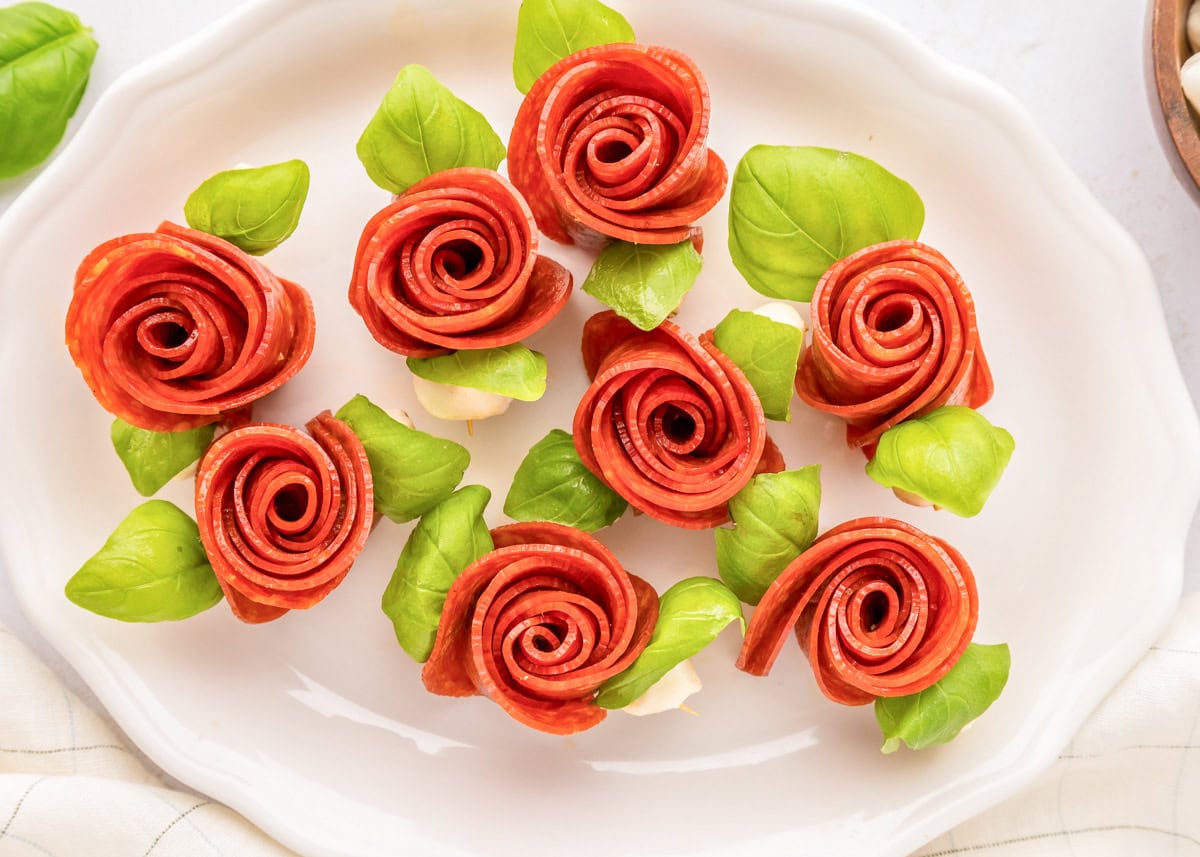 Create beautiful {and delicious} pepperoni rose skewers with only 3 ingredients and a few simple steps - a picture-perfect appetizer! Several pepperoni rose skewers on a white serving platter.