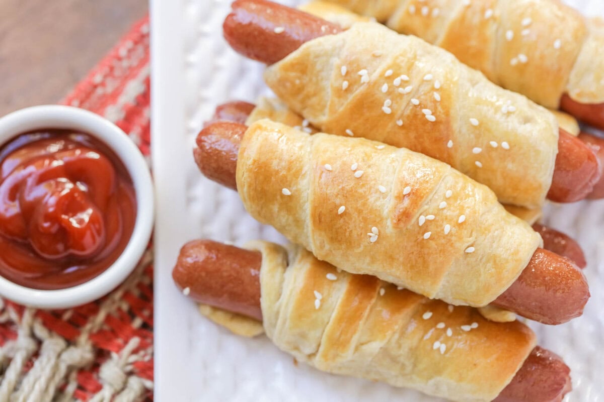 Both kids and adults will love these quick and simple pigs in a blanket. This nostalgic recipe is on the table in 20 minutes! Pigs in a Blanket on white dish served with a side of ketchup.