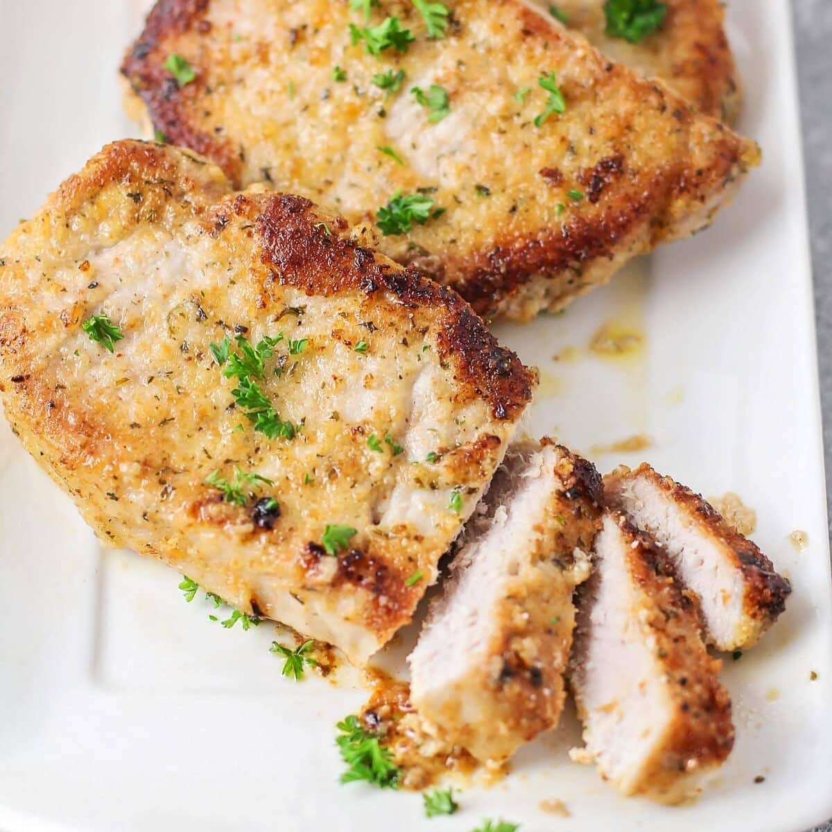 Crusted with parmesan and fried up with less than 10 minutes of prep work, this is definitely the best pork chop recipe you'll ever try!! Parmesan Crusted pork chops close up image - cut up on white plate.