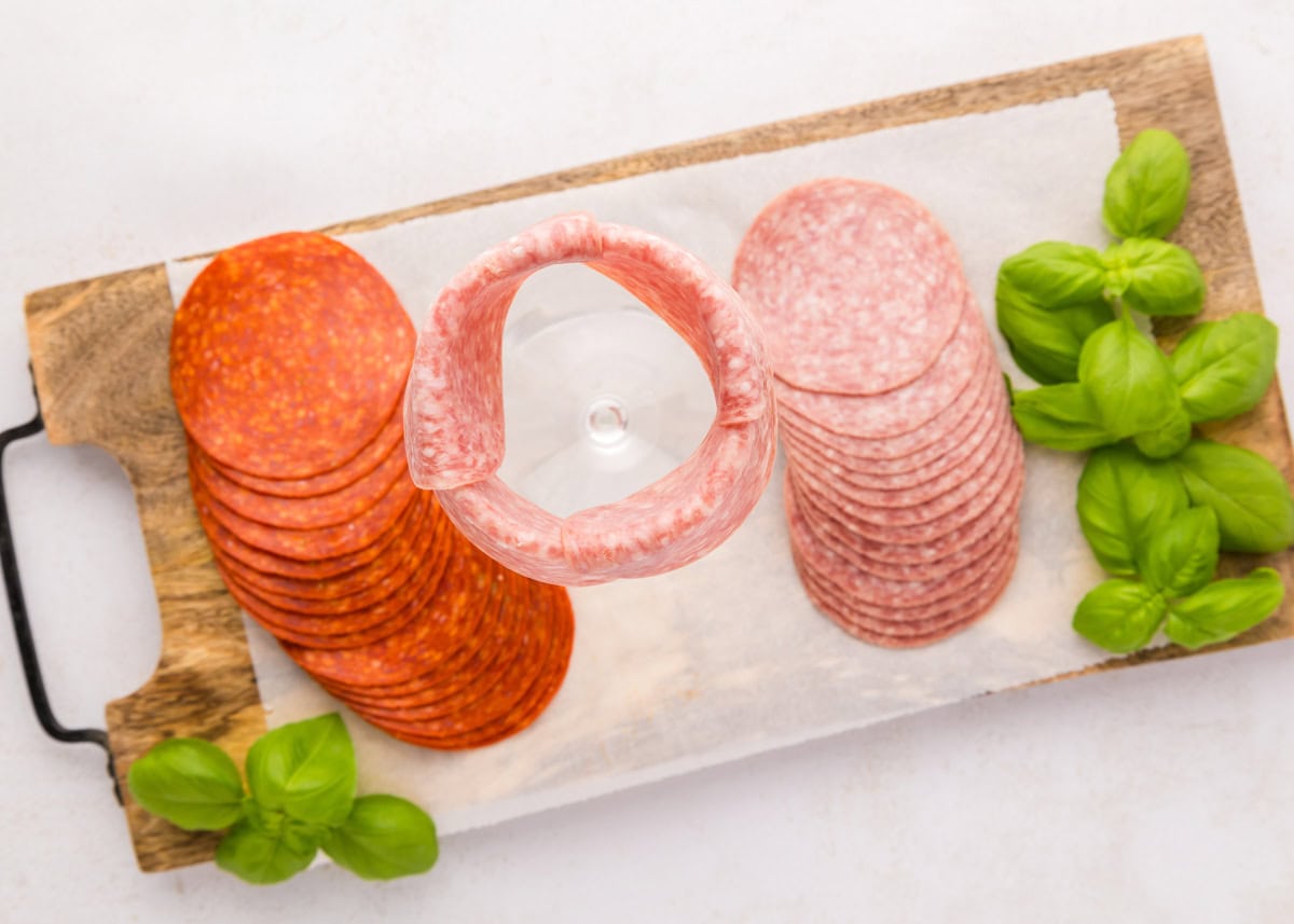 Follow these step-by-step instructions to create a beautiful salami rose - a deliciously elegant centerpiece for your snack boards! Salami Rose how to image - salami folded over the edge of a glass cup.