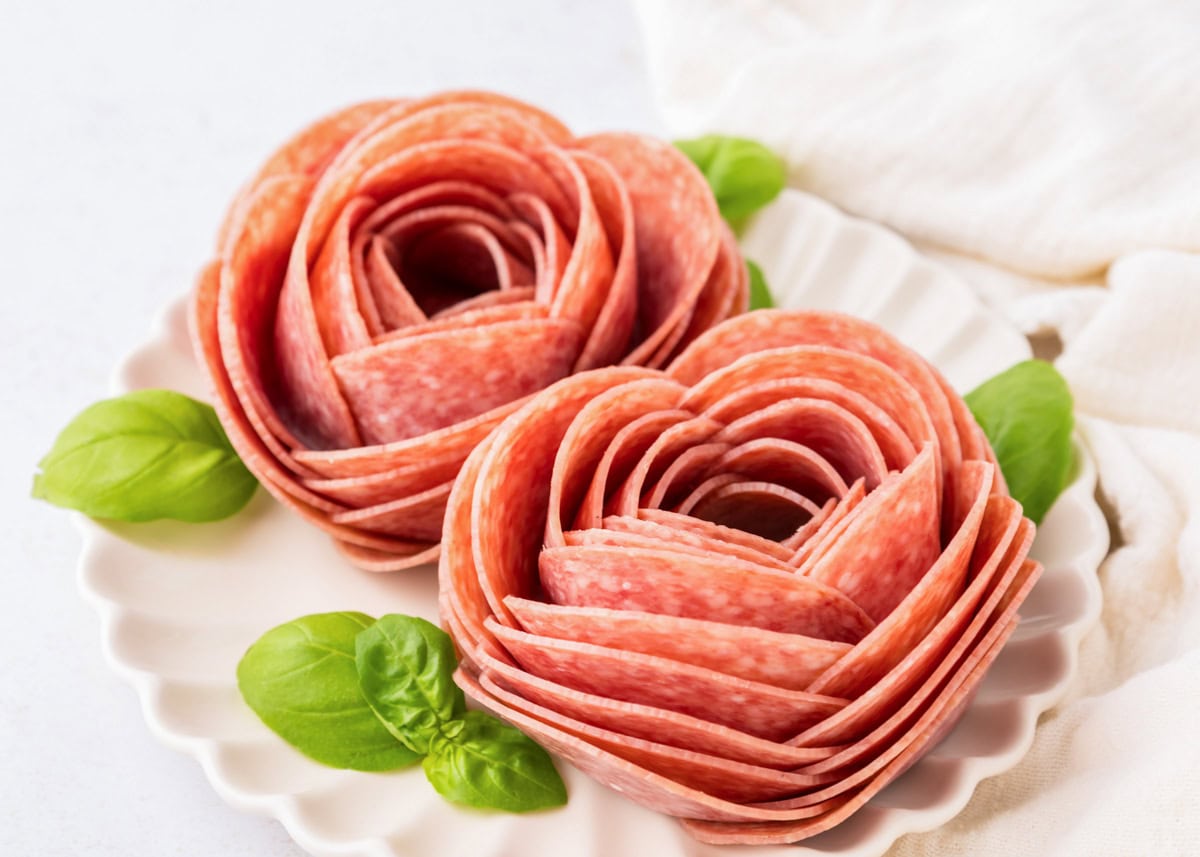 Follow these step-by-step instructions to create a beautiful salami rose - a deliciously elegant centerpiece for your snack boards! Two salami roses on a white plate.