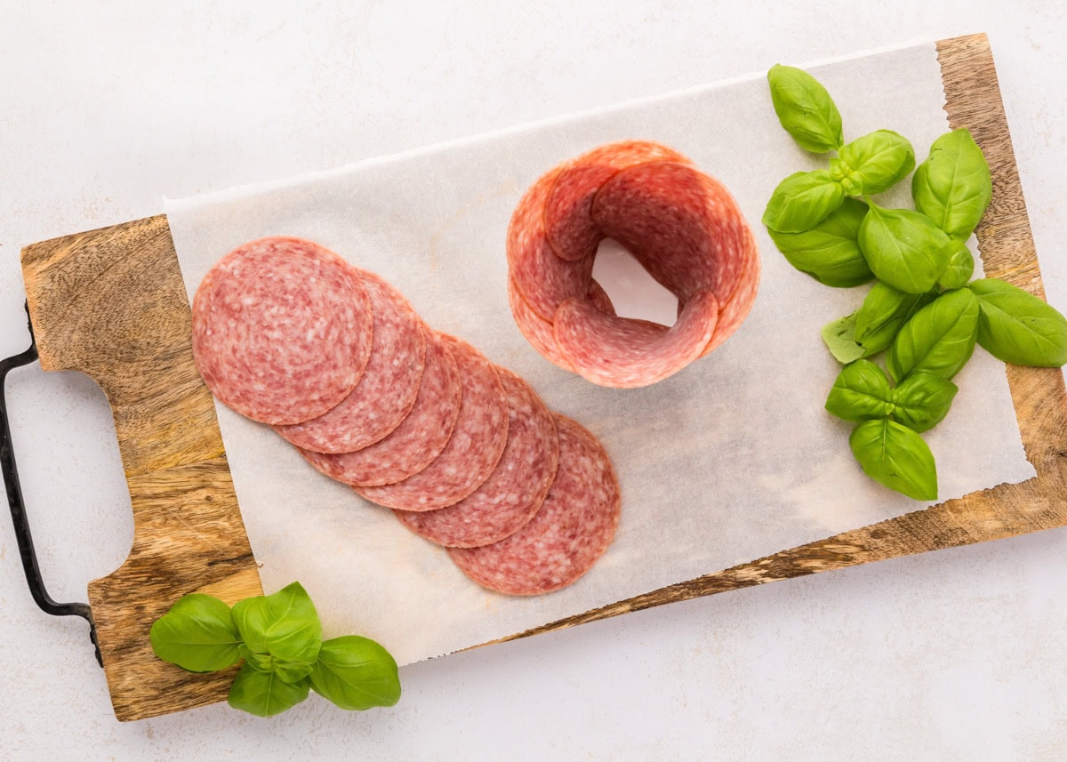 Follow these step-by-step instructions to create a beautiful salami rose - a deliciously elegant centerpiece for your snack boards! Placing layered salami around a glass jar.