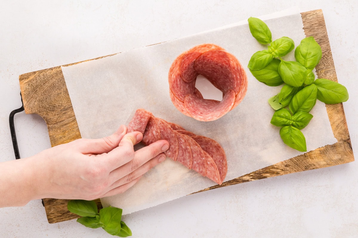Follow these step-by-step instructions to create a beautiful salami rose - a deliciously elegant centerpiece for your snack boards! Rolling layered salami on a cutting board.